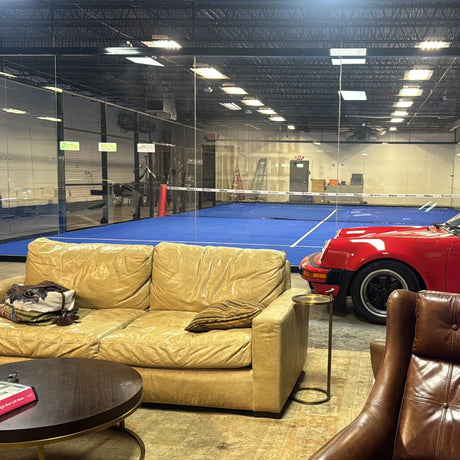 A spacious room features a yellow couch, leather chairs, and a round wooden table with books. Nearby, a red sports car is parked. Beyond the glass wall, enjoy Padel Smash’s blue padel court for a thrilling 90-min per-player rental. Exposed beams and lights adorn the ceiling.