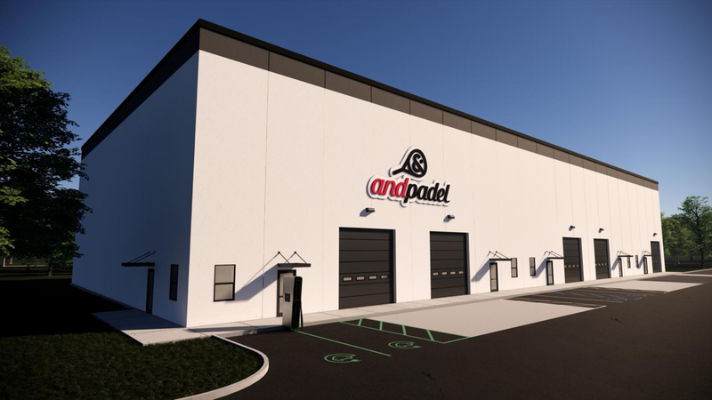 Exterior view of a modern white building with a padel logo. It has multiple garage doors and a sign above them. The surrounding area is paved with parking spaces, and trees are in the background under a clear blue sky.