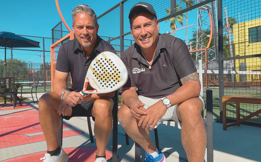 Cesc Caceres and Julian Wortelboer in black shirts sit on a bench at an outdoor padel court. One holds a padel racket. They both smile at the camera. The background shows a clear blue sky, trees, and surrounding sports facilities.
