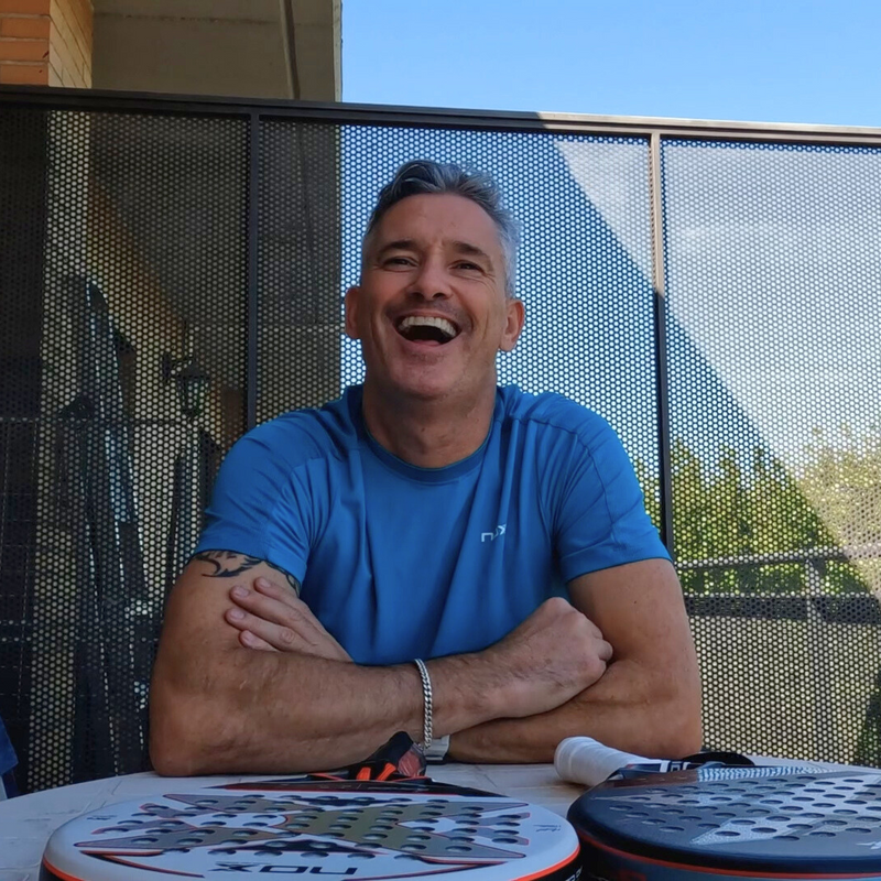 Miguel Lamperti sits at an outdoor table, smiling broadly. Hes wearing a blue shirt and has his arms crossed. Two padel rackets and a ball are on the table. Behind him is a metal mesh fence and blue sky.