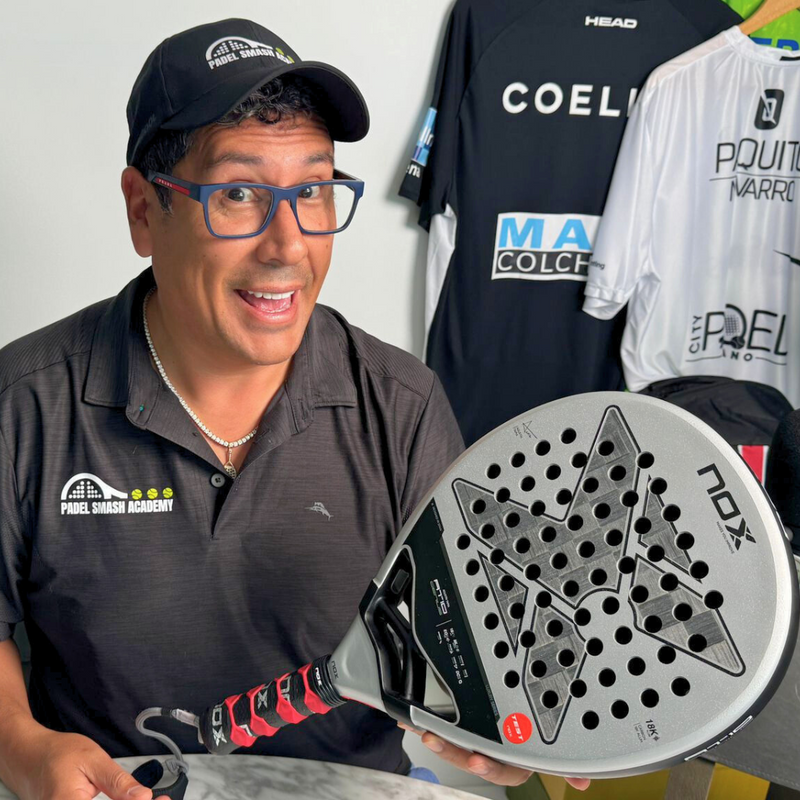 Cesc Caceres wearing a Padel Smash Academy cap and glasses holds a Nox padel racket, smiling at the camera. Behind him are padel shirts hanging on the wall with various sponsor logos. He is seated at a stone table with rackets and gear around.