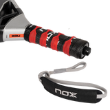Close-up of the NOX Padel Racket AT10 Genius 18K handle, wrapped in black and red grip tape. Enhanced with EOS Flap technology, it includes a gray wrist strap showing the NOX brand and features a textured, contrasting color design.