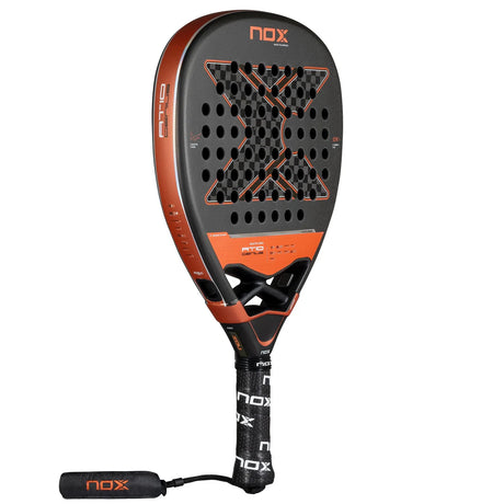 The Nox Padel Racket AT10 Genius Attack 12K 25 by Agustin Tapia features a black and orange design with a perforated X core, Exclusive Spin Technology, and is made from premium 12K Carbon. It has a black grip handle and sleek wrist strap with NOX branding.