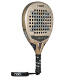 The NOX Padel Racket TL10 Future 2024 Tino Libaak features a gold and black teardrop shape with a black handle, strap, and X design. Made from aluminized 12K Carbon Fiber, it has a perforated face for versatile gameplay and a black grip with NOX printed on it.