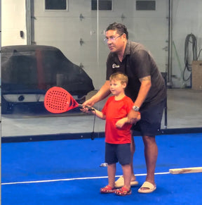 On a blue indoor sports court, a man assists a young boy holding a red paddle during a Padel Smash clinic. They focus on the swing with covered objects and other sports gear visible in the background.