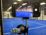 A smartphone on the Padel Smash Academys Padel Pro Mount attached to a glass pane captures the excitement of a pickleball game, highlighting dynamic movement as it tracks two players on a blue court inside a gym.